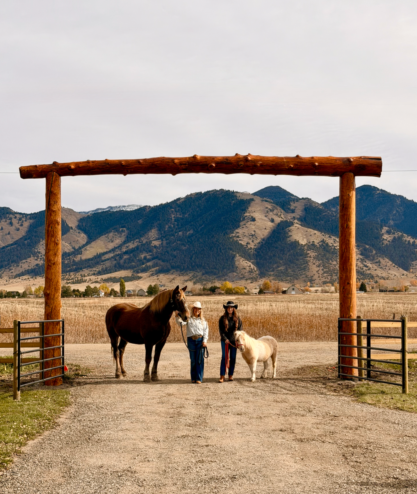 Two riders with a draft horse and miniature pony beneath a wooden ranch arch with mountains behind